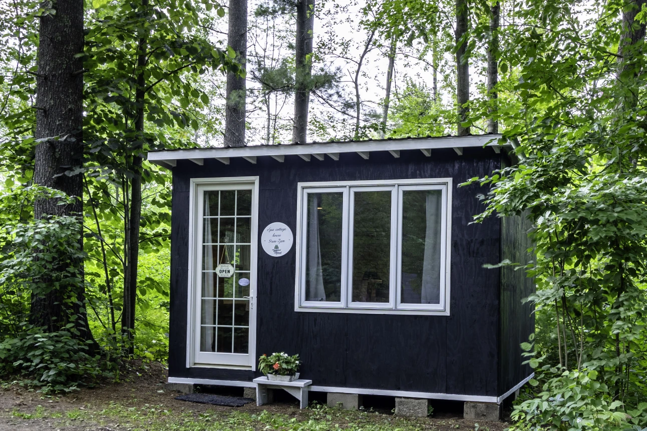 A small black cabin in the woods with a clock.